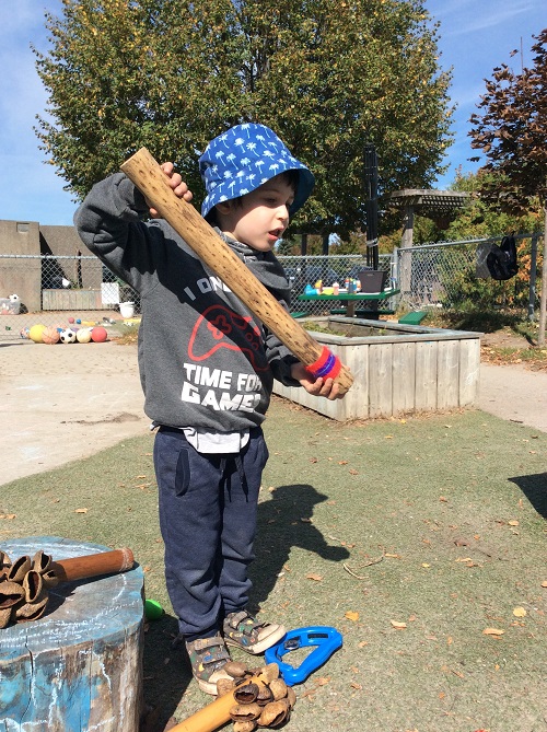Child standing on grass, holding a large wooden percussion instrument.