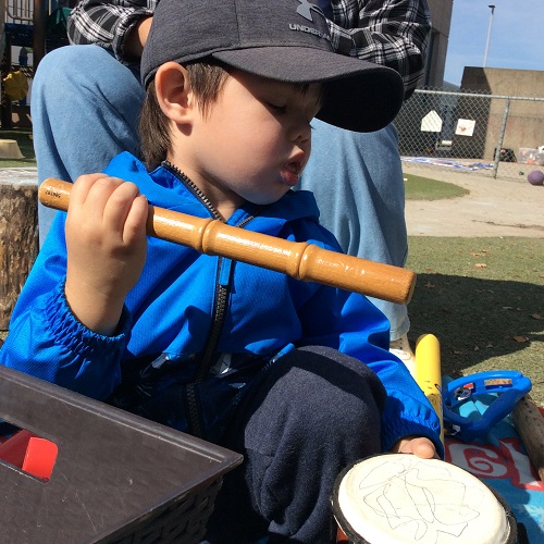 Little boy in blue jacket sitting on the ground, striking small drum with wooden stick.