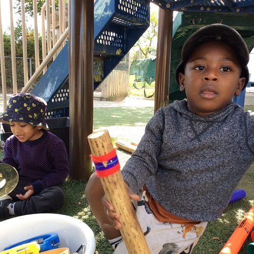 Two children sitting under a playground structure holding musical instruments.