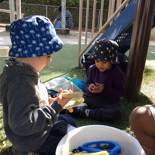 Two children sit on turf near a bucket of instruments, playing with cymbal and shaker.