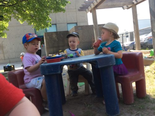 Three children sitting at plastic outdoor table engaged in pretend play, using toy food and dishes.