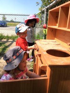 Children playing with a wooden outdoor play kitchen, pretending to cook using the stove and oven.