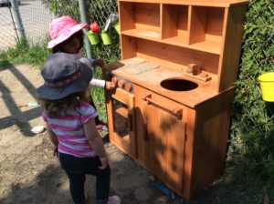 Two children standing in front of the play kitchen, discussing and mimicking cooking activities.