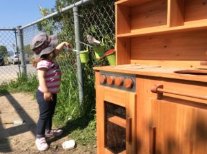 Child using a ladle to scoop into green hanging buckets beside the wooden play kitchen outdoors.