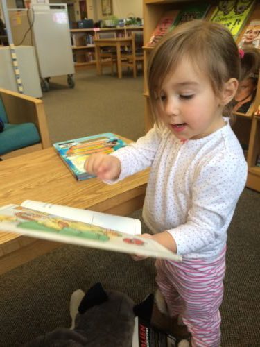 Young girl holding a book and showing a picture while reading aloud.