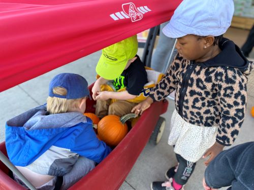 Children in wagon with pumpkins, with a little girl standin beside wagon.