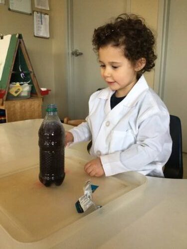 Child wearing white lab coat observing a bottle filled with dark liquid during science experiment.