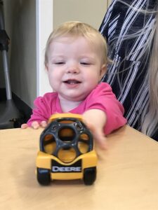 Infant enthusiastically reaching for a yellow toy truck while sitting at a table.