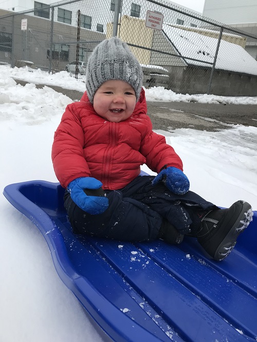 Infant bundled in red jacket and grey toque smiling while sitting on a blue sled in the snow.