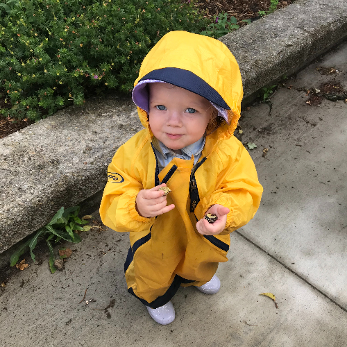 Child in a yellow rain suit standing on wet pavement looking up.