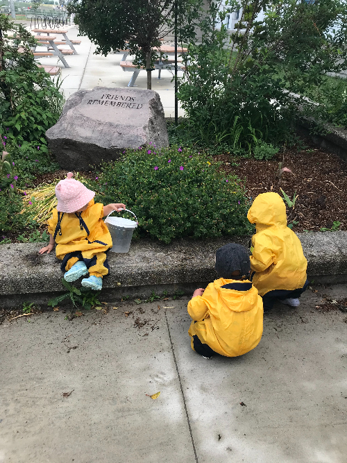 Three children in yellow rain suits playing near large stone that reads “Friends Remembered.”