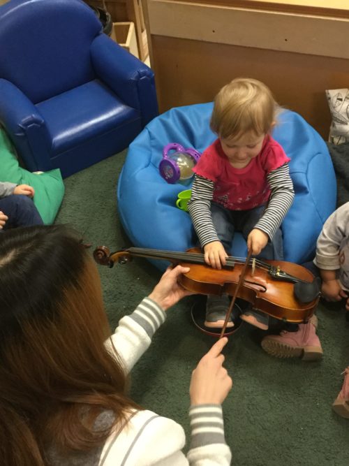 A toddler smiles while seated on a bean bag, using a bow to play a violin held by an adult.