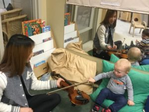 A baby reaches out to touch the violin bow held by visiting Seneca College student.