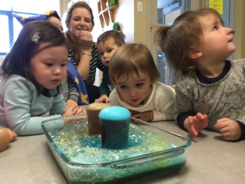 Children gathered around a clear dish, watching colourful foam erupt from volcano experiment.