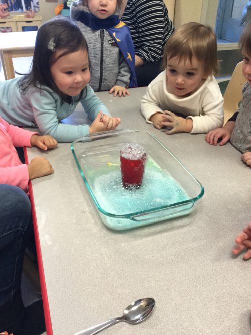 Children observing red foam bubbling up from a cup placed in a glass baking dish.