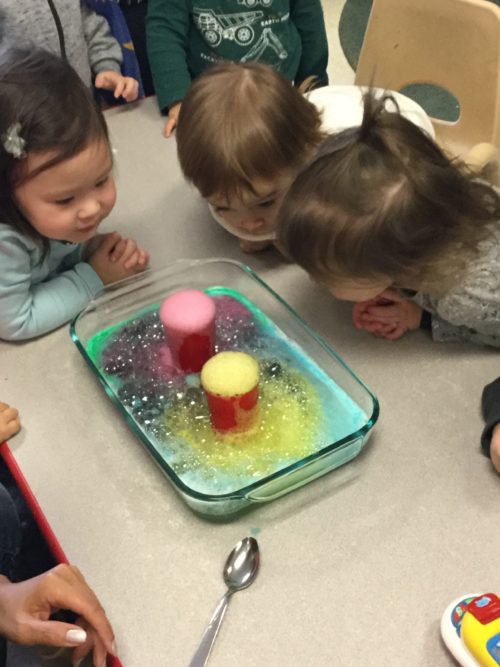 Three toddlers leaning in to watch a vibrant, foaming reaction during volcano science activity.