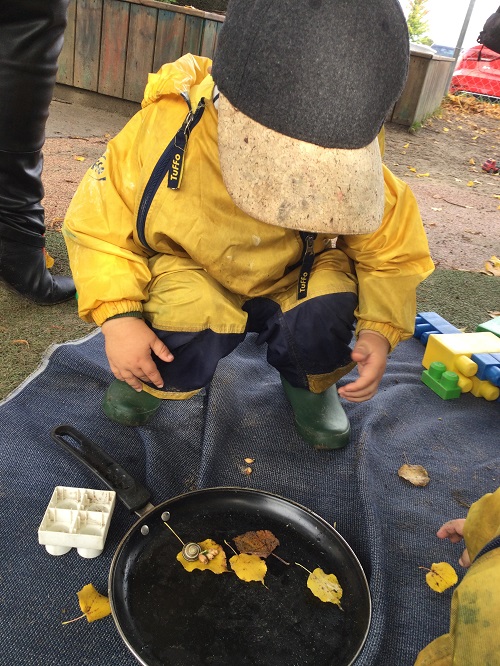 Child in yellow coat on blanket, crouched over frying pan with snail and autumn leaves.