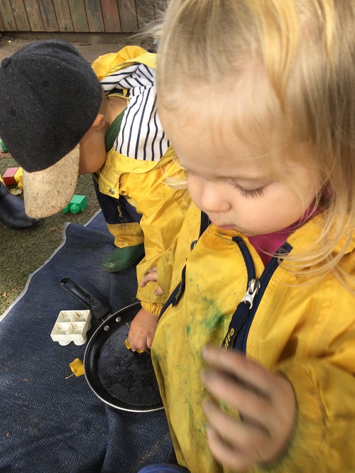 Two children in yellow raincoats outside playing with frying pan.
