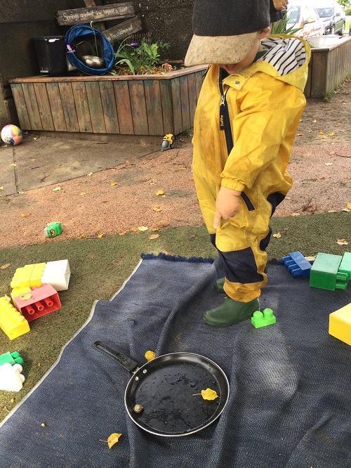 Child standing outside on blanket in yellow raincoat looking down at a frying pan.
