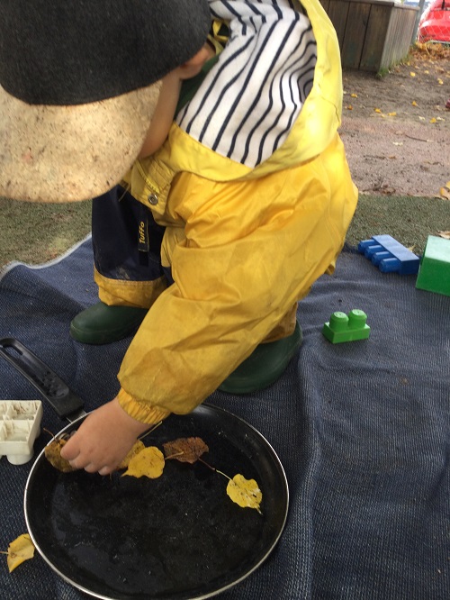 Child in a yellow raincoat placing leaves in frying pan beside snail.