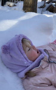 Child wearing purple beanie laying down on the snow, closing their eyes