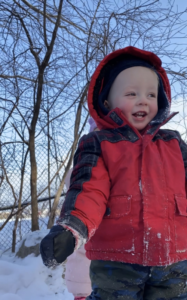 Happy toddler boy in red jacket and hood standing outside in winter with bare trees behind him