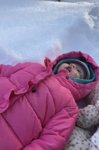 Child wearing pink jacket and hood laying down on the snow